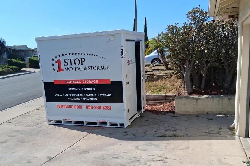 A 1-Box container standing on a residential driveway beside a house, one door open. Its side still carries One Stop Moving & Storage lettering.