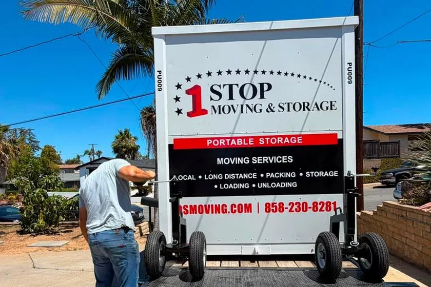 A 1-Box container being loaded onto a tilt trailer in a driveway with the ramp down, a worker standing at the side of the trailer.